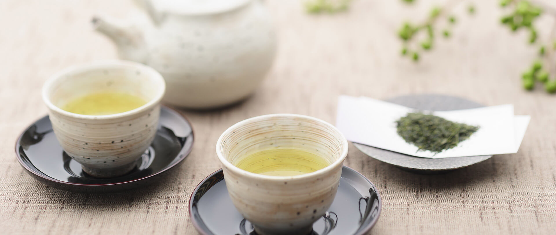 Two cups of green tea sit side-by-side. A teapot and a shallow bowl of ground green tea leaves lie in the background.