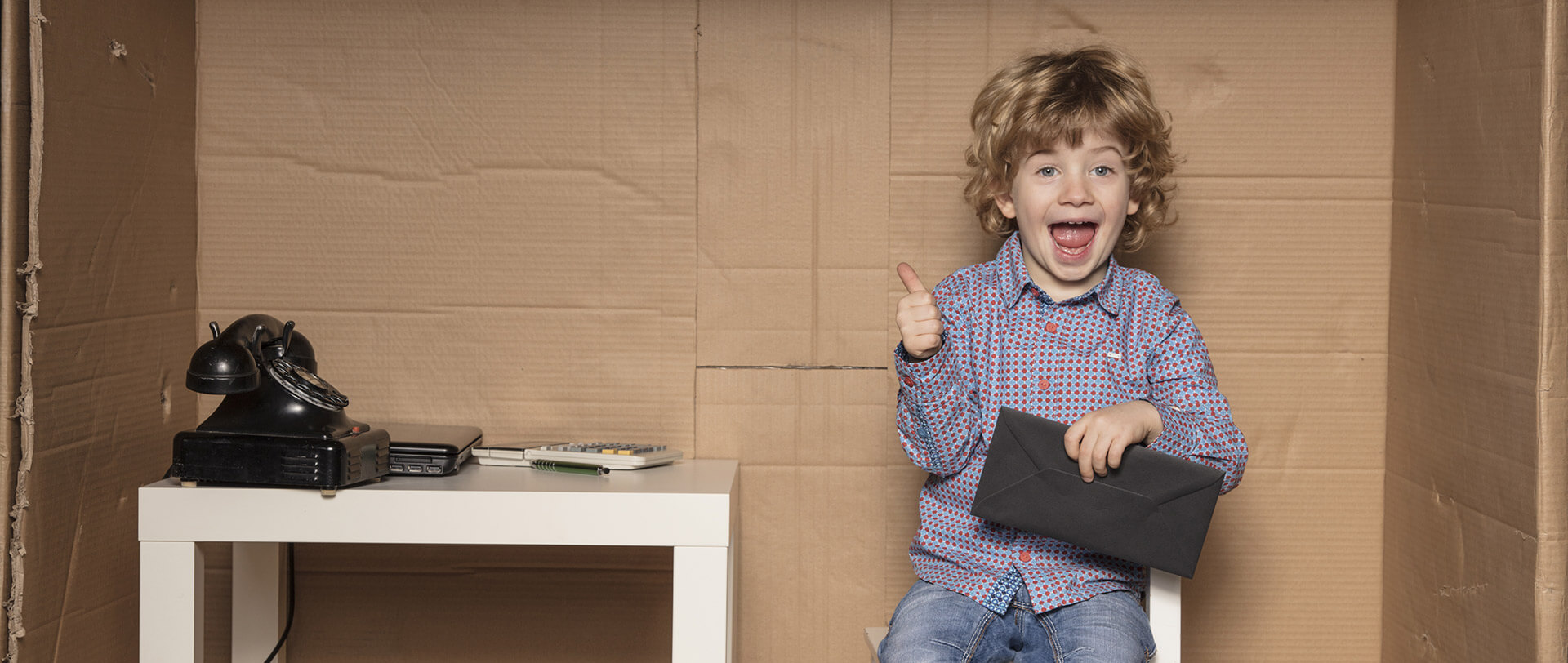 • A boy in business-casual clothing sits in a kid-sized cardboard office cubicle and flashes a grin and a thumbs-up